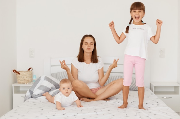 Free photo portrait of young adult other doing morning exercises in yoga pose while her little daughters playing near mommy, preschooler girl standing with clenched fists, toddler lying on tummy.