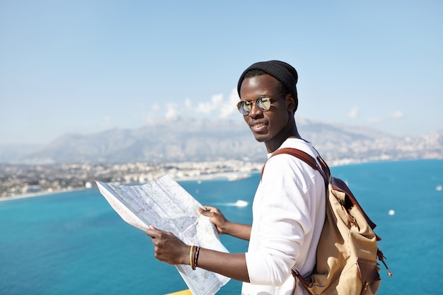 Portrait of young African American traveler looking with paper map in his hands, wearing sunglasses and hat, standing on viewing platform, admiring European city and beautiful seascape