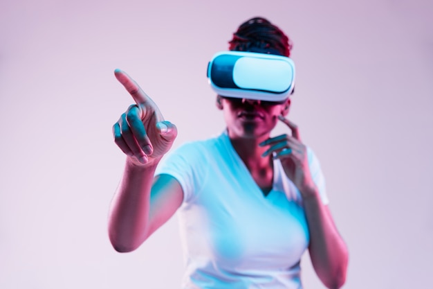 Portrait of young african-american woman's playing in VR-glasses in neon light on gradient background