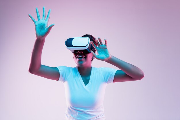 Portrait of young african-american woman's playing in VR-glasses in neon light on gradient background