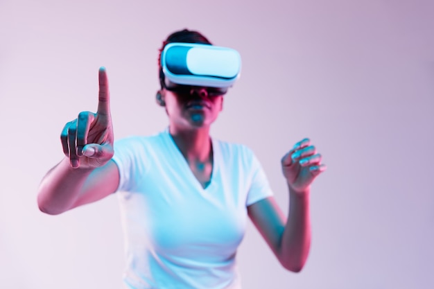 Portrait of young african-american woman's playing in VR-glasses in neon light on gradient.