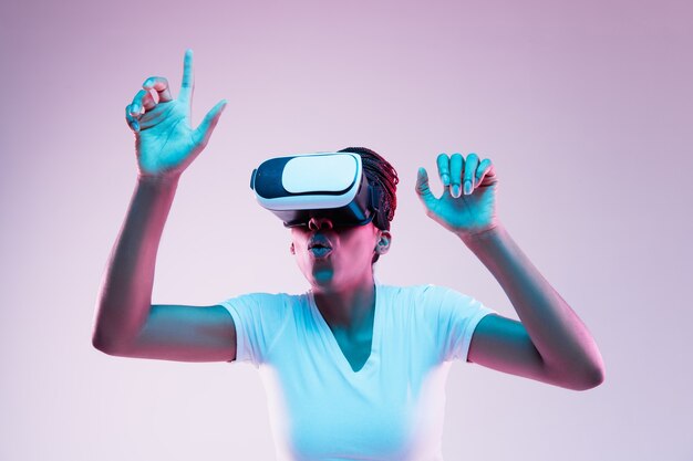 Portrait of young african-american woman's playing in VR-glasses in neon light on gradient