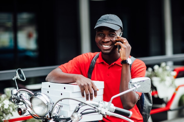 Free Photo portrait of young african guy accepts the order by phone in motorbike holding boxes with pizza and sit on his bike. urban place.