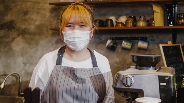 Portrait young Asia girl waitress wear medical face mask feeling happy smile waiting for clients after lockdown at urban cafe.