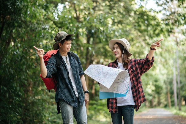 Portrait Young asian handsome man with backpack and trekking hat and pretty girlfriend standing and checking direction on paper map while walking on forest trail, backpack travel concept