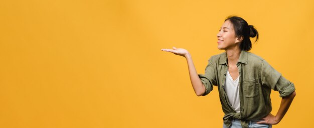 Portrait of young Asian lady smiling with cheerful expression, shows something amazing at blank space in casual clothing and standing isolated over yellow wall. Panoramic banner with copy space.