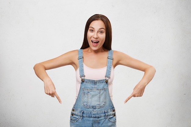 Portrait of young attractive brunette female wearing white T-shirt and denim overalls, pointing with fore fingers down, having happy expression while advertising something. Woman showing something