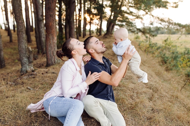 Free photo portrait of young attractive family with little baby son, posing in beautiful autumn pine forest at sunny day. handsome man and  his pretty brunette wife