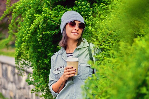 Free Photo portrait of young attractive female in sunglasses holds paper coffee cup in a green summer park.