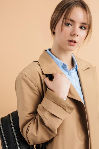 Portrait of young attractive girl in trench coat and blue shirt with backpack on shoulder while dreamily looking in camera over beige background