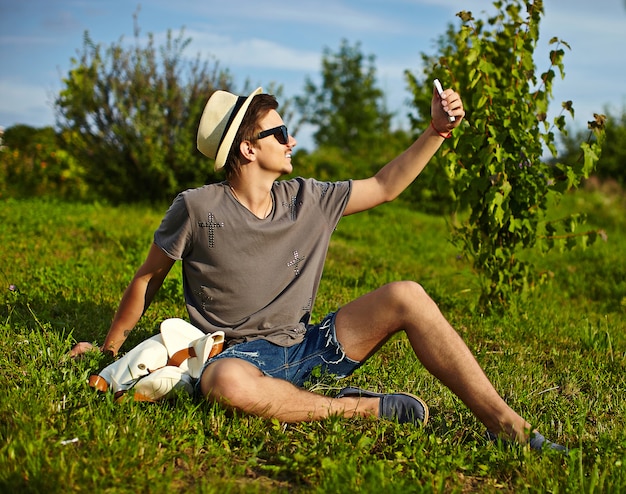 portrait of young attractive modern stylish man in casual cloth in hat in glasses sitting in the park in green grass making selfie