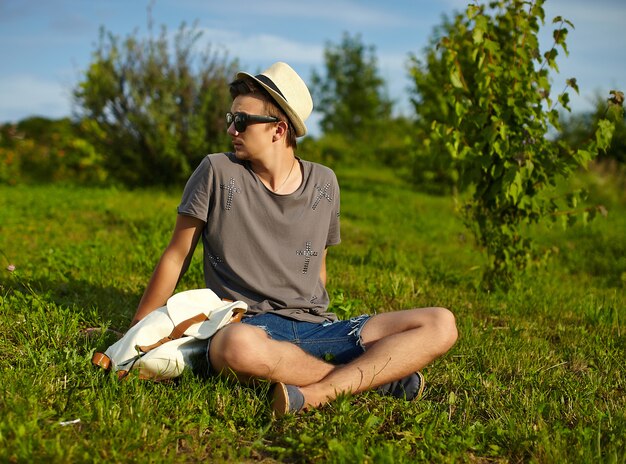 portrait of young attractive modern stylish man in casual cloth in hat in glasses sitting in the park in green grass