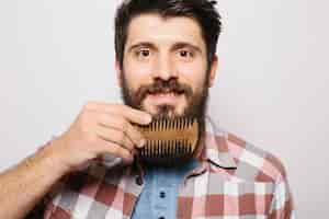 Free photo portrait of young attractive redhead hipster male with serious and confident look, holding wooden comb and doing his thick beard. stylish bearded barber in checkered shirt combing in salon. horizontal