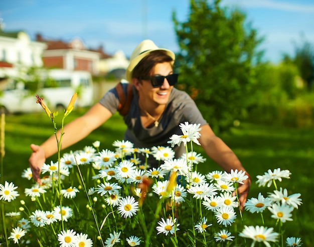 portrait of young attractive smiling modern stylish man in casual cloth in hat in glasses in the park with bright colorful flowers in camomiles