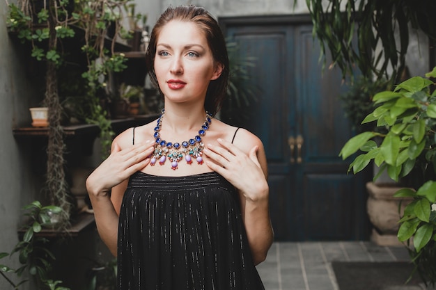 Portrait of young attractive woman in elegant black dress wearing luxury rich necklace jewelry, summer style, fashion trend, vacation, stylish accessories, posing on tropical villa on Bali