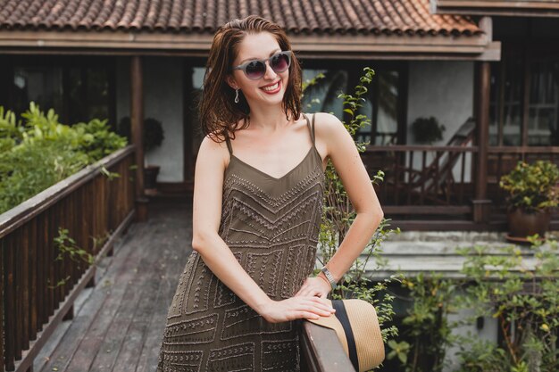 Portrait of young attractive woman in elegant dress, straw hat, summer style, fashion trend, vacation, smiling, stylish accessories, sunglasses, posing on tropical villa on Bali