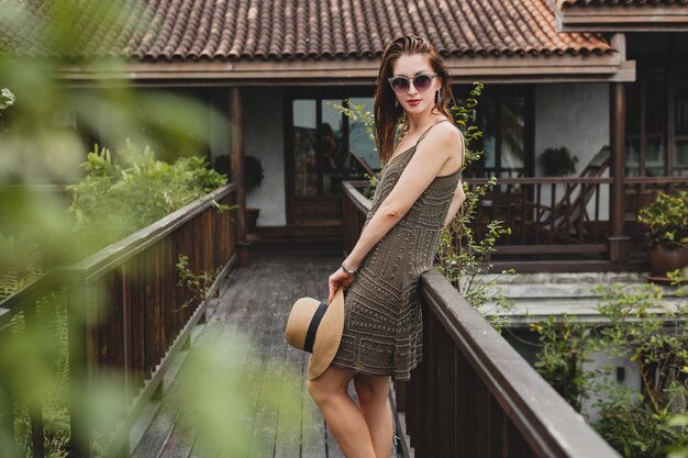 Portrait of young attractive woman in elegant dress, straw hat, summer style, fashion trend, vacation, smiling, stylish accessories, sunglasses, posing on tropical villa on Bali