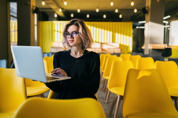 Portrait of young attractive woman sitting in lecture hall working on laptop wearing glasses, student learning in classroom with many yellow chairs