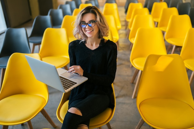 Portrait of young attractive woman sitting in lecture hall working on laptop wearing glasses, student learning in classroom with many yellow chairs