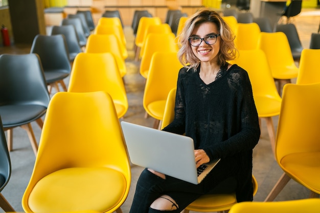 Portrait of young attractive woman sitting in lecture hall working on laptop wearing glasses, student learning in classroom with many yellow chairs