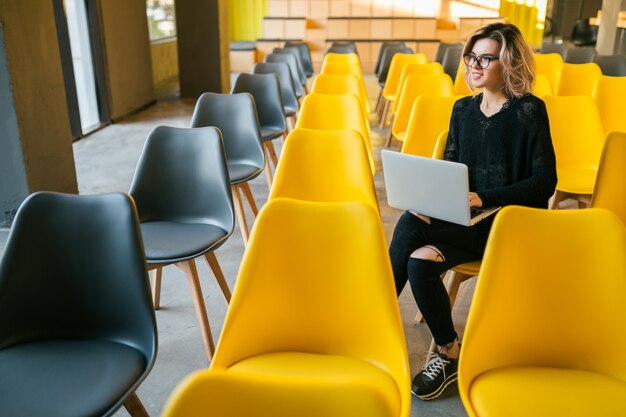 Portrait of young attractive woman sitting in lecture hall working on laptop wearing glasses, student learning in classroom with many yellow chairs