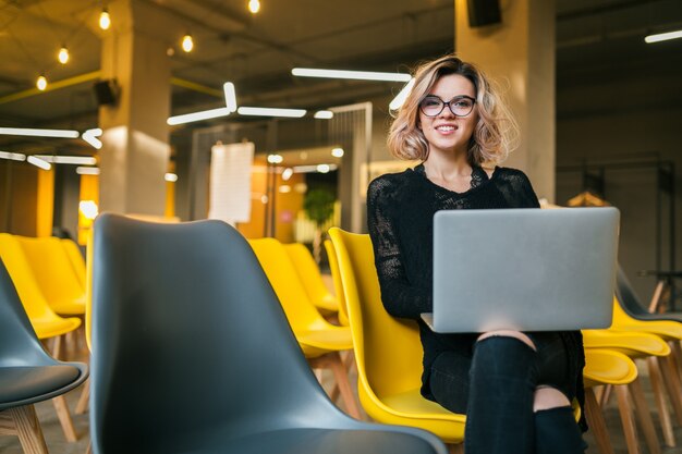 Portrait of young attractive woman sitting in lecture hall working on laptop wearing glasses, student learning in classroom with many yellow chairs