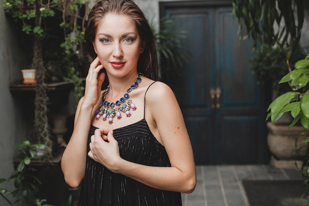 Portrait of young attractive woman in stylish black dress posing at tropical villa, sexy, elegant summer style, fashionable necklace accessories, smiling, jewelry, pure natural skin of face