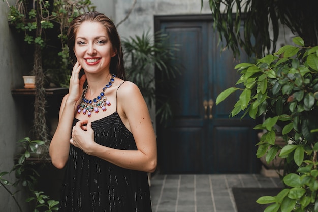 Portrait of young attractive woman in stylish black dress posing at tropical villa, sexy, elegant summer style, fashionable necklace accessories, smiling, jewelry, pure natural skin of face