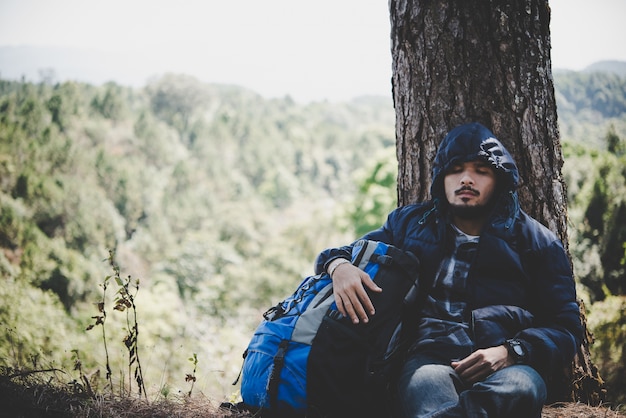 Free photo portrait of young beard man sitting alone by a tree with backpack looking away enjoy with nature.