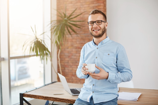 Free photo portrait of young bearded entrepreneur in glasses and casual clothes, standing in bright coworking office