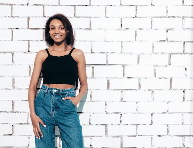 Free photo portrait of young beautiful black woman smiling model dressed in summer jeans and black top clothes sexy carefree female posing near white brick wall in studio tanned and cheerful