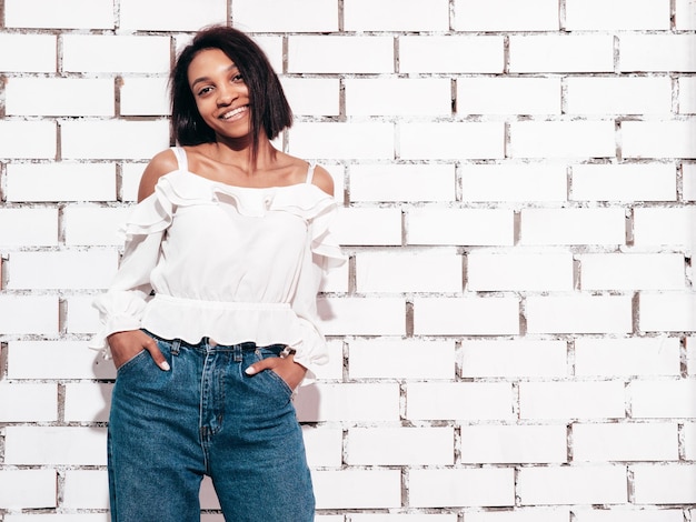 Free Photo portrait of young beautiful black woman smiling model dressed in summer jeans clothes sexy carefree female posing near white brick wall in studio tanned and cheerful