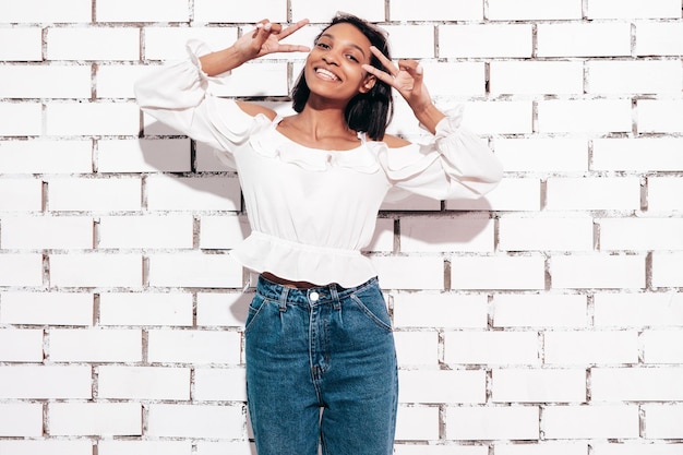 Free Photo portrait of young beautiful black woman smiling model dressed in summer jeans clothes sexy carefree female posing near white brick wall in studio tanned and cheerful