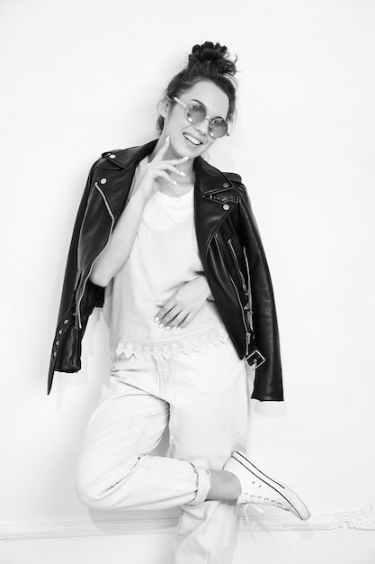 Portrait of young beautiful brunette woman girl model with nude makeup wearing summer hipster biker leather jacket clothes in sunglasses posing near wall. 