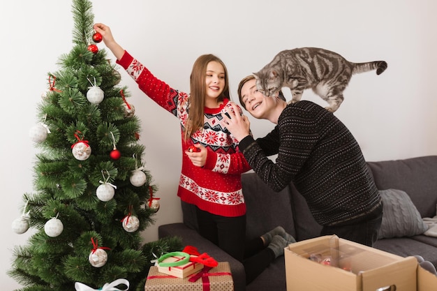 Portrait of young beautiful couple with cat standing at home and decorating Christmas tree together