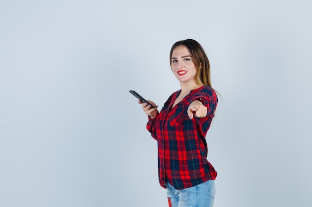 Portrait of young beautiful female holding phone, pointing at front in casual shirt and looking glad front view