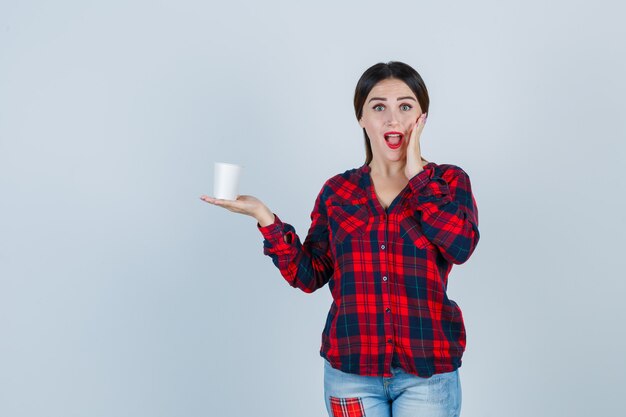 Portrait of young beautiful female holding plastic glass, keeping hand on face in casual shirt, jeans and looking perplexed front view