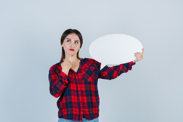 Portrait of young beautiful female holding speech bubble while keeping finger on chin in casual shirt, jeans and looking thoughtful front view