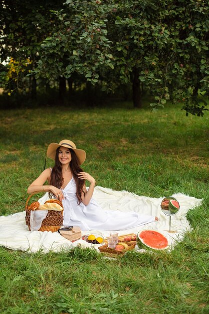 Portrait of a young beautiful girl with even white teeth, a beautiful smile in a straw hat and long white dress have a picnic in the garden