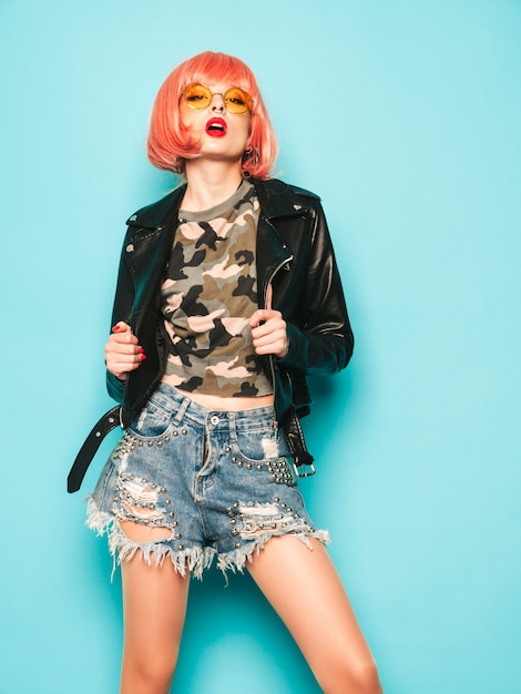 Portrait of young beautiful hipster bad girl in black leather jacket and earring in her nose.Sexy carefree smiling woman sitting in studio in pink wig near blue wall.Positive model having fun