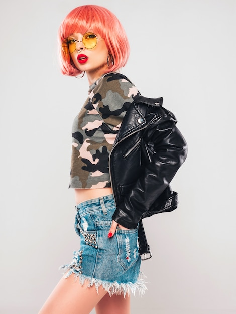 Portrait of young beautiful hipster bad girl in black leather jacket and earring in her nose.Sexy carefree smiling woman sitting in studio in pink wig near gray wall.Confident model in sunglasses