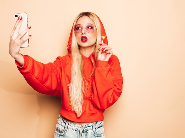 Portrait of young beautiful hipster bad girl in trendy jeans  clothes and earring in her nose.Sexy carefree smiling blond woman takes selfie.Positive model licking round sugar candy