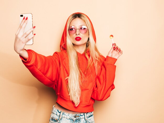 Portrait of young beautiful hipster bad girl in trendy jeans  clothes and earring in her nose.Sexy carefree smiling blond woman takes selfie.Positive model licking round sugar candy