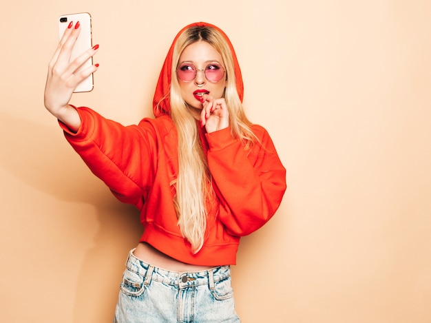 Portrait of young beautiful hipster bad girl in trendy jeans  clothes and earring in her nose.Sexy carefree smiling blond woman takes selfie.Positive model licking round sugar candy