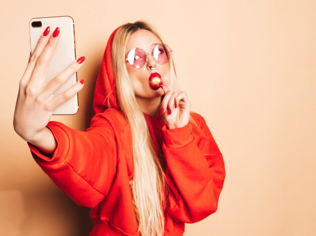 Portrait of young beautiful hipster bad girl in trendy jeans  clothes and earring in her nose.Sexy carefree smiling blond woman takes selfie.Positive model licking round sugar candy