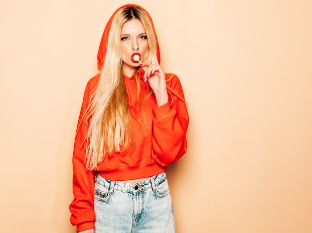 Free Photo portrait of young beautiful hipster bad girl in trendy red  hoodie and earring in her nose.  positive model licking round sugar candy