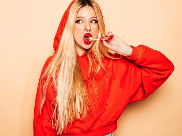 Free photo portrait of young beautiful hipster bad girl in trendy red  hoodie and earring in her nose.  positive model licking round sugar candy