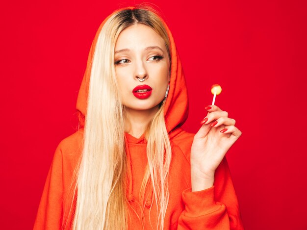 Portrait of young beautiful hipster bad girl in trendy red  hoodie and earring in her nose.  Positive model licking round sugar candy