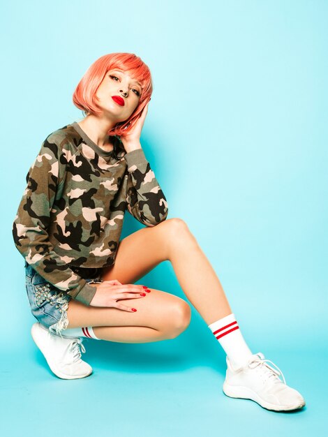 Portrait of young beautiful hipster bad girl in trendy red summer clothes and earring in her nose.Sexy carefree smiling woman sitting in studio in pink wig near blue wall.Positive model having fun