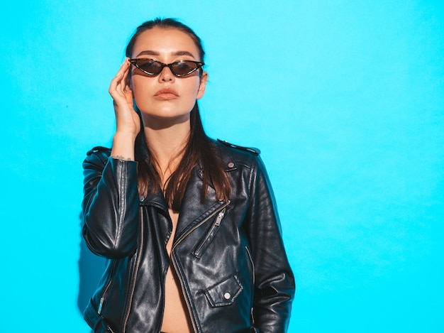 Portrait of young beautiful hipster  bad girl in trendy summer black leather jacket and sunglasses. Sexy carefree woman isolated on blue. Brunette model posing